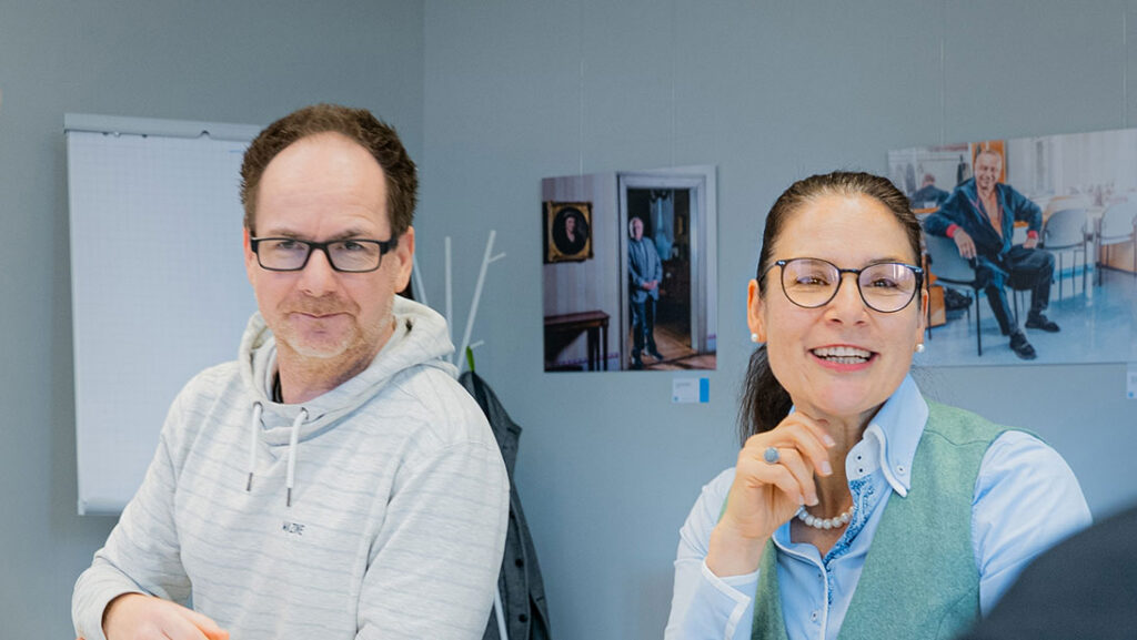 Zwei Personen sitzen an einem Tisch in einem Raum mit Fotografien an der Wand.
