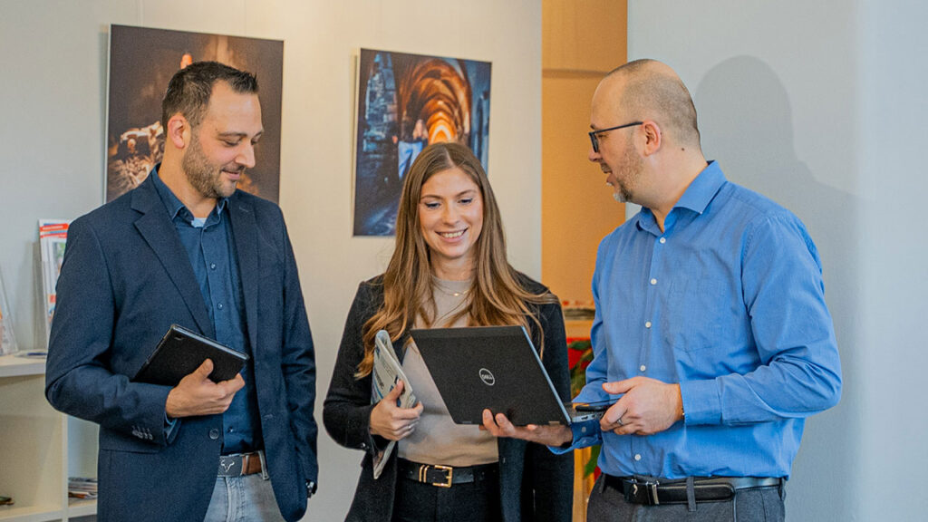 Drei Personen stehen zusammen und betrachten einen Laptop in einem Innenraum.