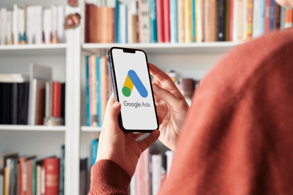 Eine Person hält ein Smartphone in der Hand. Auf dem Bildschirm ist das Logo von Google Ads zu sehen. Im Hintergrund befindet sich ein Bücherregal mit zahlreichen Büchern. ©AdobeStock