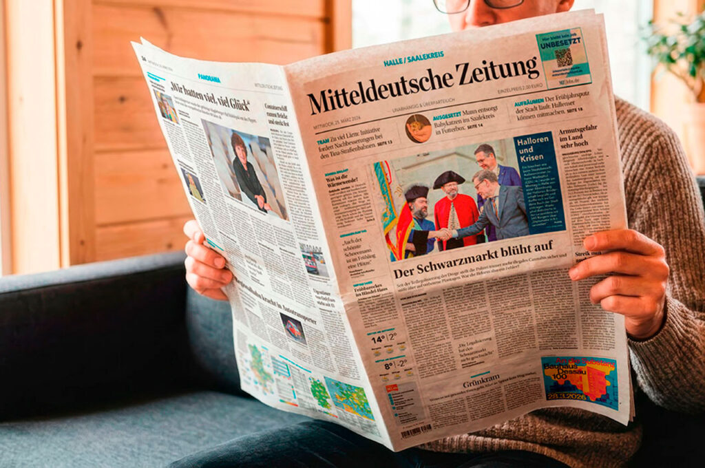 Person sitzt auf einem Sofa und liest eine gedruckte Tageszeitung. Die Titelseite der Zeitung ist sichtbar, während der Wohnraum im Hintergrund unscharf dargestellt ist. ©Mockup AdobeStock, Inhalt Mediengruppe Mitteldeutschland