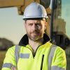 Fachkraft auf einer Baustelle mit weißem Schutzhelm, Warnschutzjacke und Baumaschine im Hintergrund ©AdobeStock